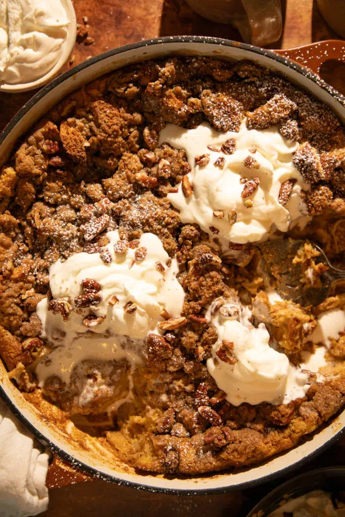Pumpkin brioche French toast bake with pecans, partially scooped, showing soft custardy inside topped with whipped cream and streusel.