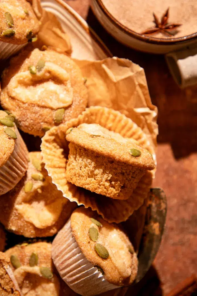 Apple cider donut muffin with liner peeled away, showing tender spiced crumb and cinnamon sugar pepita topping.