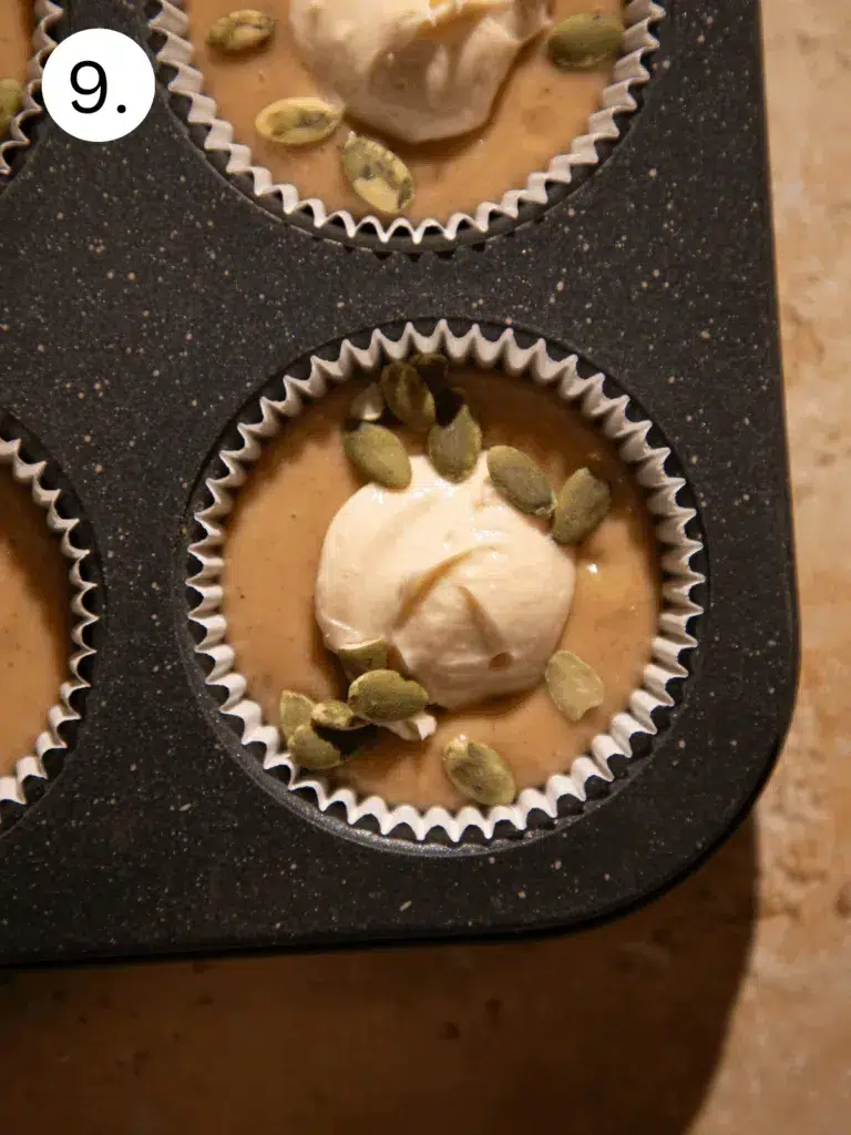 Muffin cups filled with apple cider batter, a scoop of cream cheese filling, and sprinkled pepitas before baking.