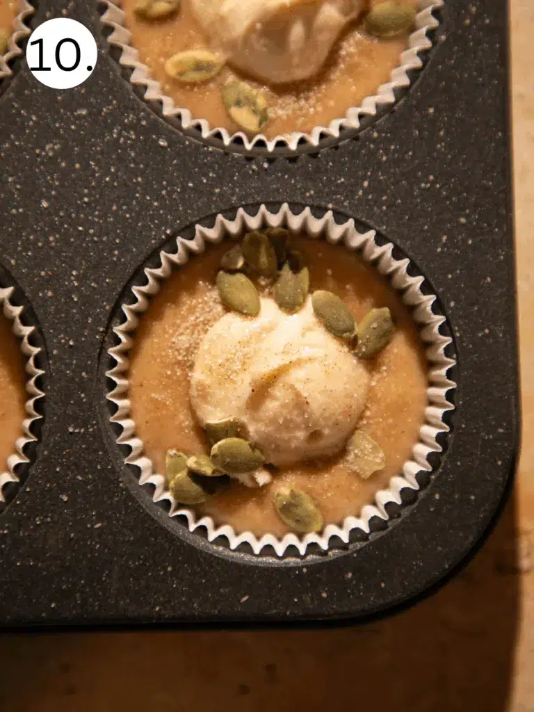 Close-up of muffin tin with apple cider donut muffin batter topped with cream cheese and pepitas.