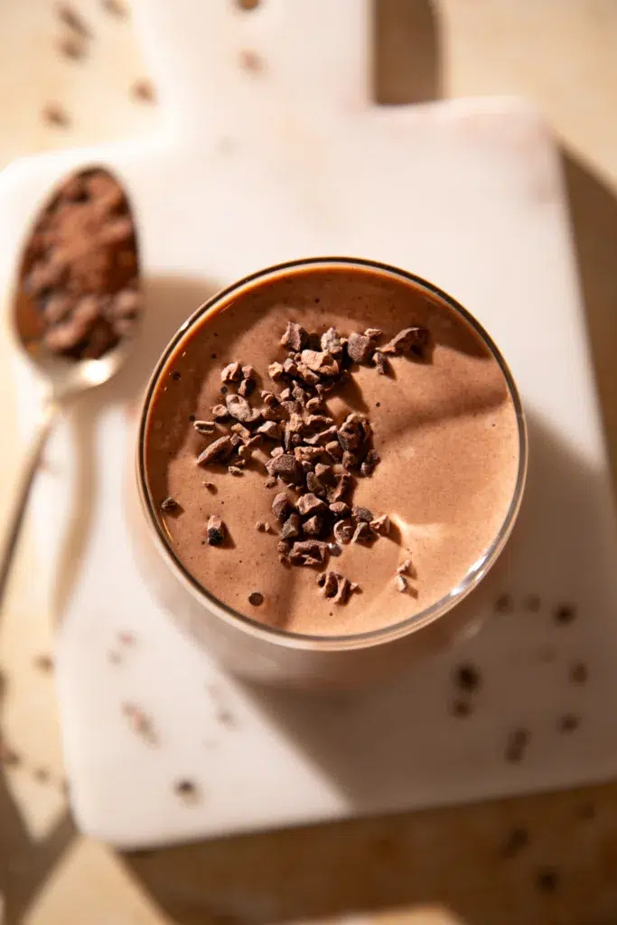 Top view of a chocolate cherry espresso protein shake in a glass, sprinkled with crunchy cacao nibs on a white board