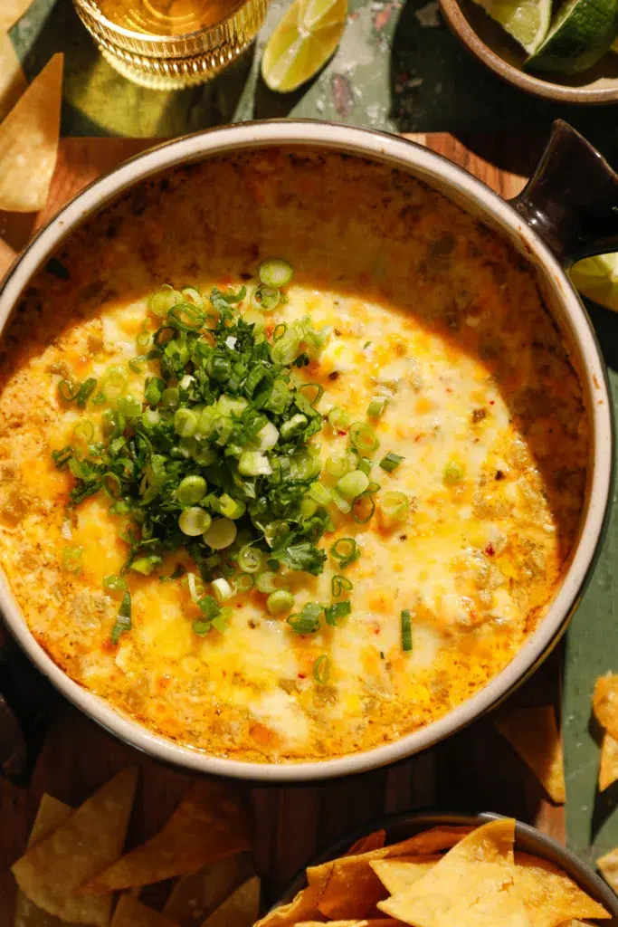 Hot cheesy baked green chile dip topped with chopped green onions in a round baking dish with tortilla chips.