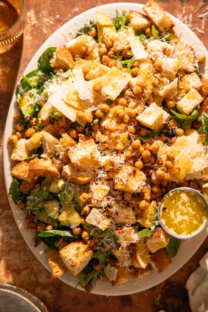 Large platter of spinach Caesar salad topped with roasted chickpeas, avocado chunks, sourdough croutons, parmesan shavings, and homemade Caesar dressing.