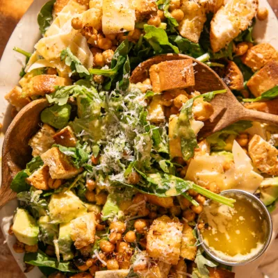 Spinach Caesar salad on platter with wooden serving spoons, topped with avocado, parmesan, and roasted chickpeas.