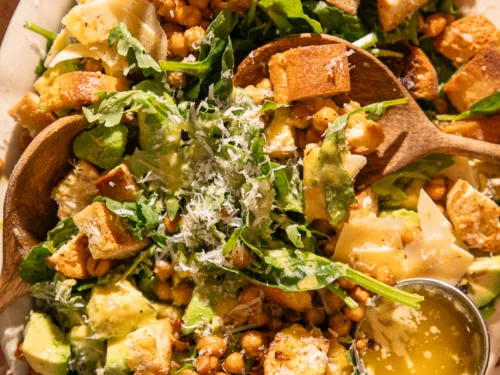 Spinach Caesar salad on platter with wooden serving spoons, topped with avocado, parmesan, and roasted chickpeas.