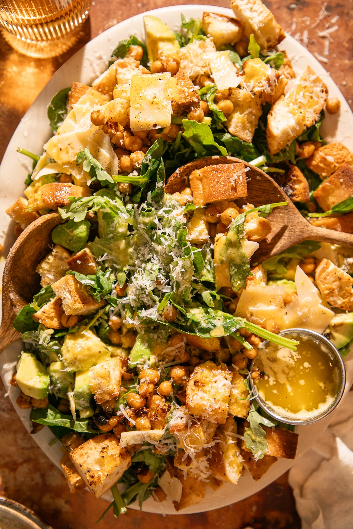 Spinach Caesar salad on platter with wooden serving spoons, topped with avocado, parmesan, and roasted chickpeas.