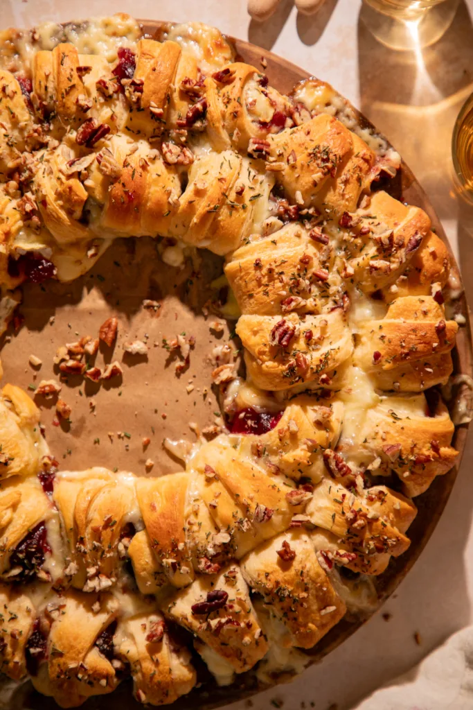 Golden baked crescent ring filled with cranberry sauce and brie cheese, topped with pecans and rosemary, served on a baking stone.