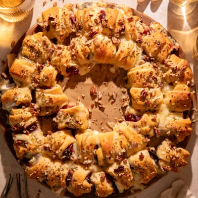 Festive cranberry brie crescent ring on a round baking stone surrounded by drinks and serving forks, perfect for entertaining.