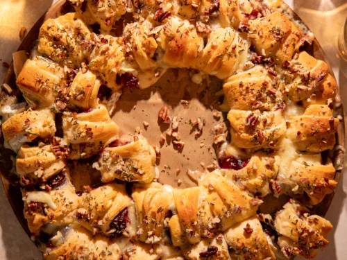 Festive cranberry brie crescent ring on a round baking stone surrounded by drinks and serving forks, perfect for entertaining.