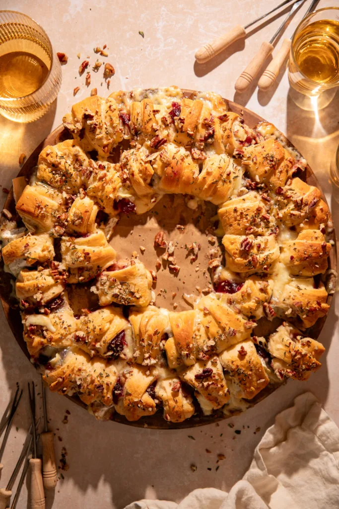 Festive cranberry brie crescent ring on a round baking stone surrounded by drinks and serving forks, perfect for entertaining.