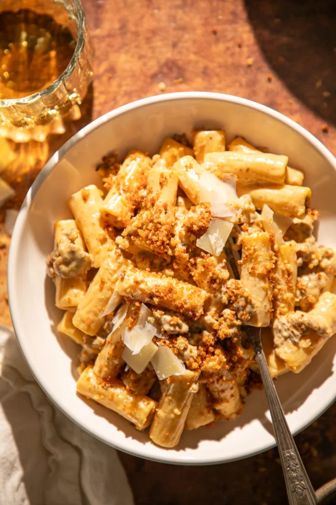 Bowl of rigatoni with pumpkin Alfredo sauce and sausage, topped with toasted breadcrumbs and Parmesan, with a fork twirled in the pasta.