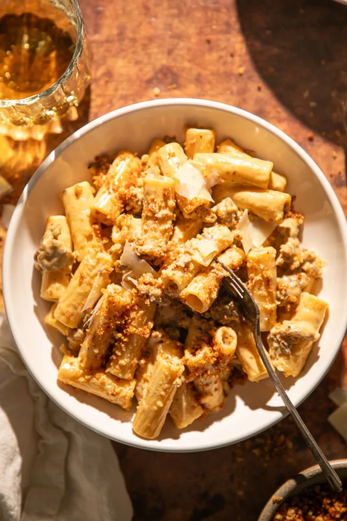 Warm-toned shot of creamy pumpkin Alfredo rigatoni served in a white bowl with golden breadcrumb topping and a glass of wine beside it.