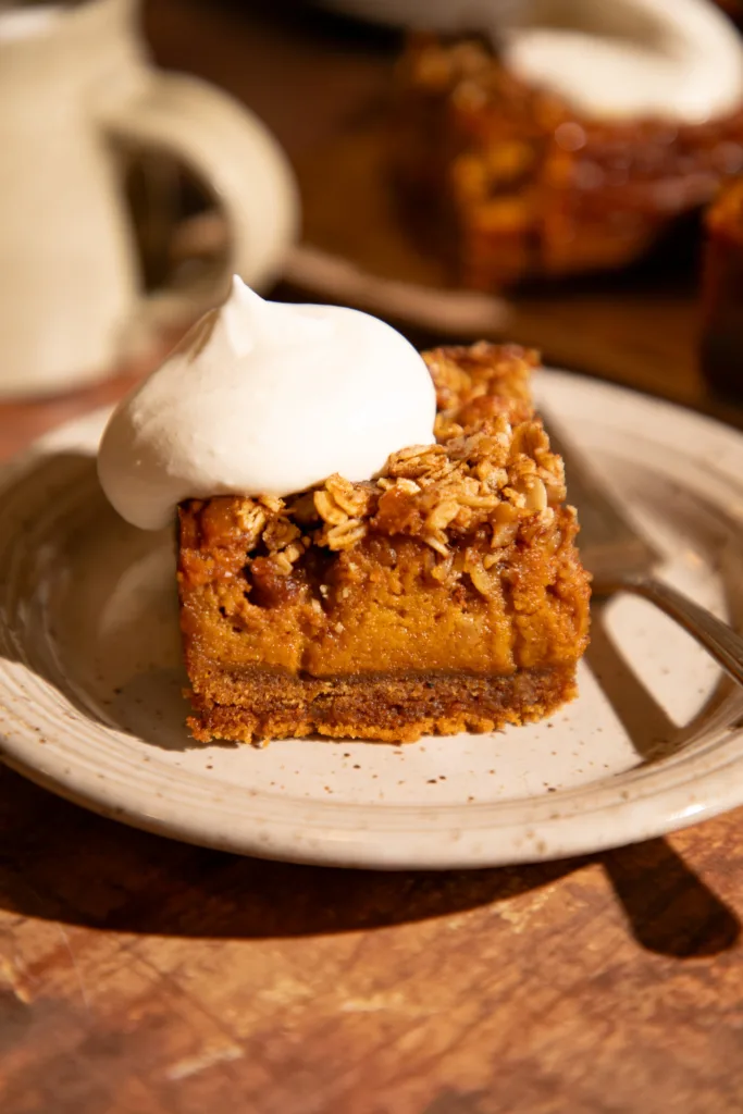 Slice of pumpkin pie bar with graham cracker crust and oat streusel topping, served on a plate with whipped cream.