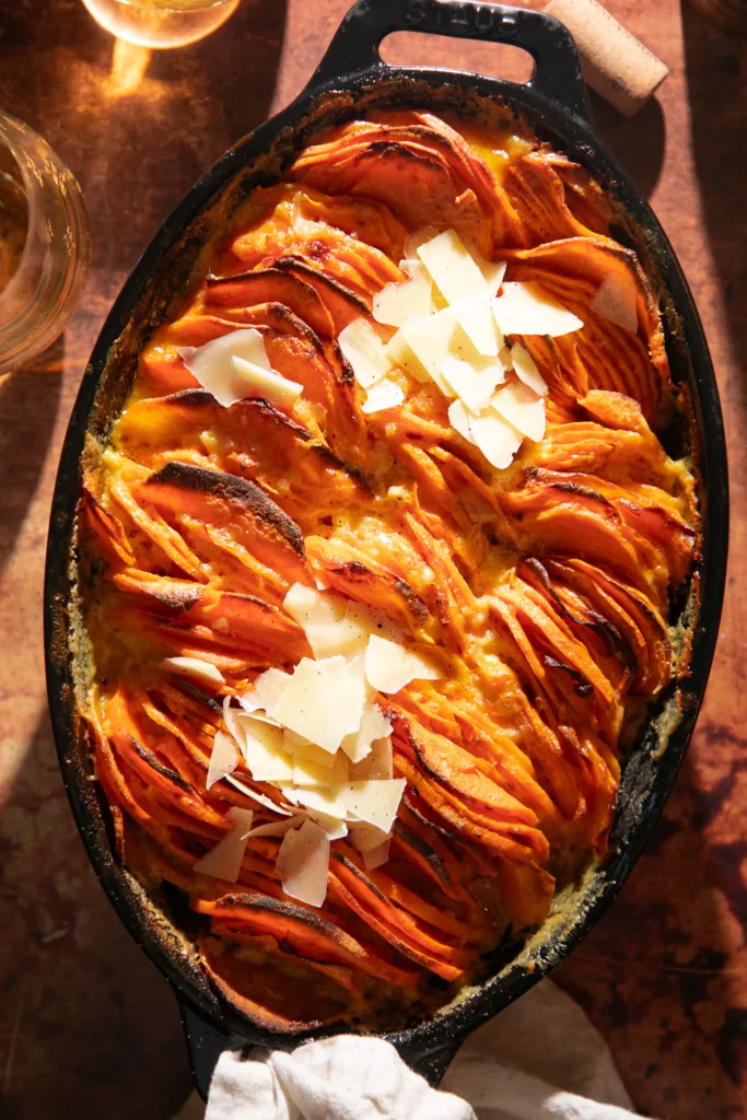 Warm-toned overhead shot of sweet potato gratin in a black oval Staub dish, topped with melted parmesan and served with white wine.