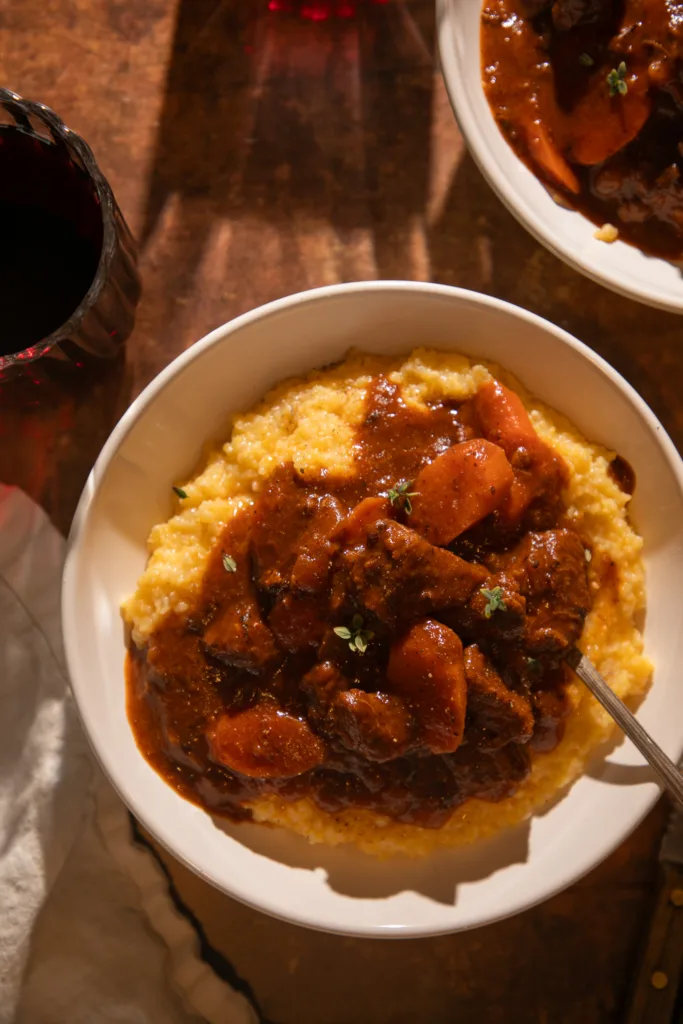 Warm, angled shot of a bowl filled with tender red wine braised beef tips and carrots served over creamy polenta, garnished with thyme.