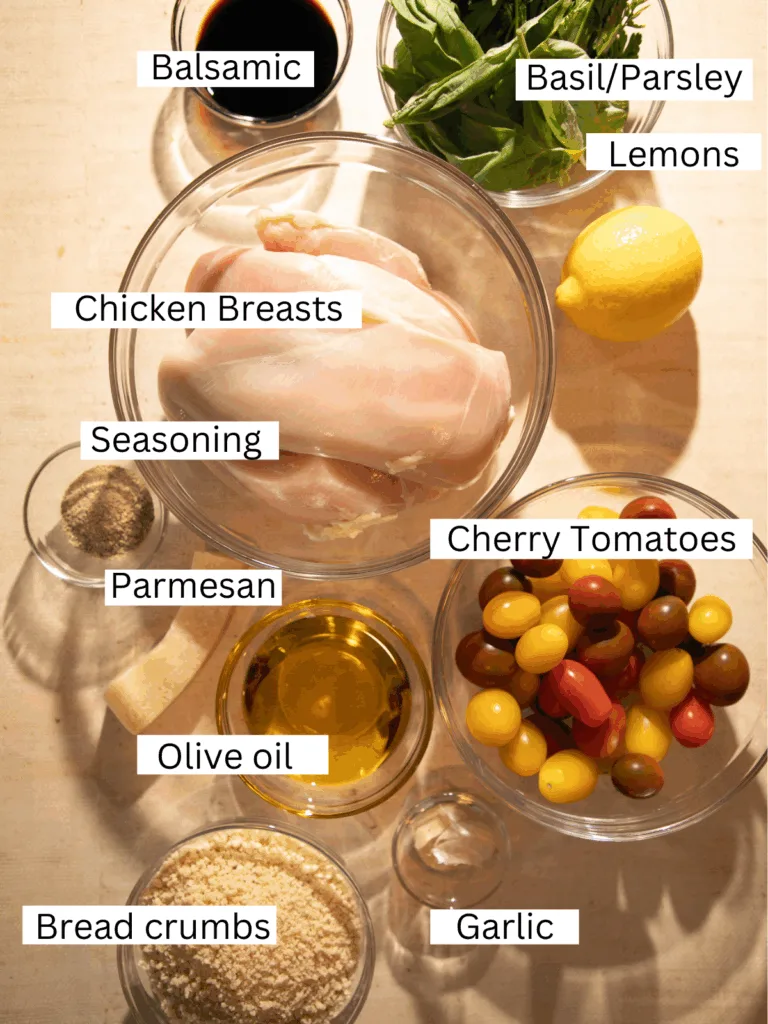Overhead view of ingredients for crispy lemon herb bruschetta chicken including chicken breasts, cherry tomatoes, basil, parsley, lemon, garlic, Parmesan, and breadcrumbs on a warm-toned surface.