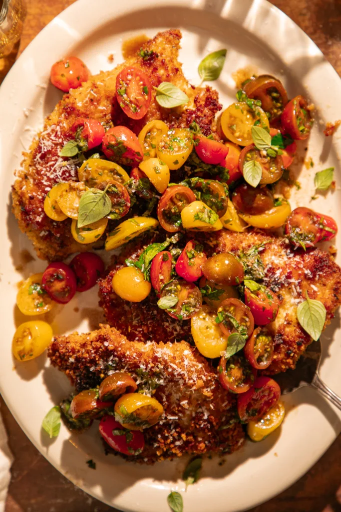 Golden, breaded chicken cutlets topped with fresh lemon herb bruschetta made from colorful cherry tomatoes and herbs, served family-style on a rustic table.