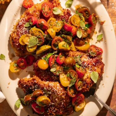Crispy chicken cutlets topped with lemon herb tomato bruschetta made with cherry tomatoes, basil, and parsley, served on a white platter with grated Parmesan and olive oil drizzle.