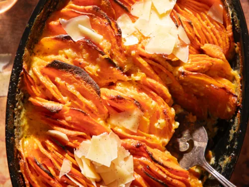 Overhead view of baked sweet potato gratin topped with shaved parmesan in a black cast iron skillet on a rustic brown surface.