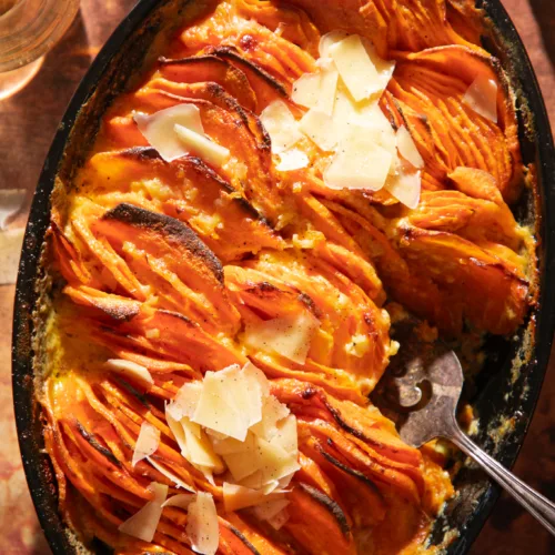 Overhead view of baked sweet potato gratin topped with shaved parmesan in a black cast iron skillet on a rustic brown surface.