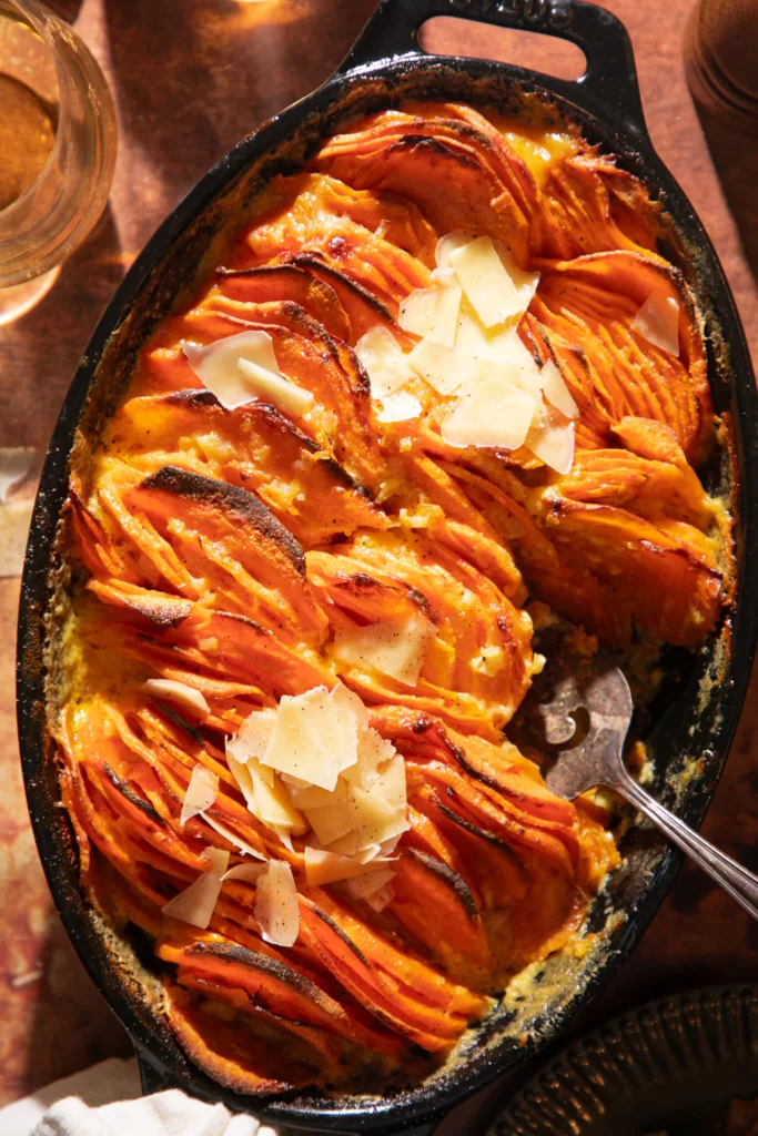 Overhead view of baked sweet potato gratin topped with shaved parmesan in a black cast iron skillet on a rustic brown surface.