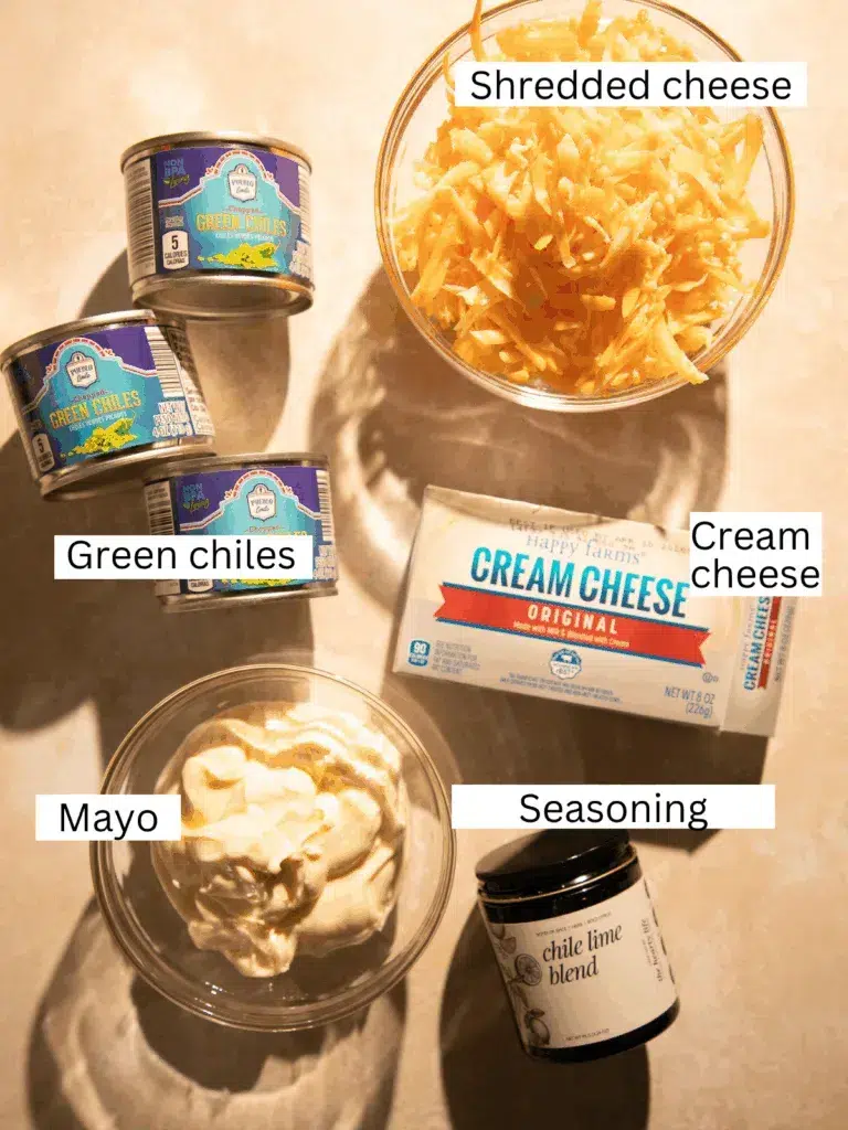 Overhead photo of recipe ingredients for cheesy baked green chile dip including canned green chiles, shredded cheddar cheese, cream cheese, mayonnaise, and seasoning blend.
