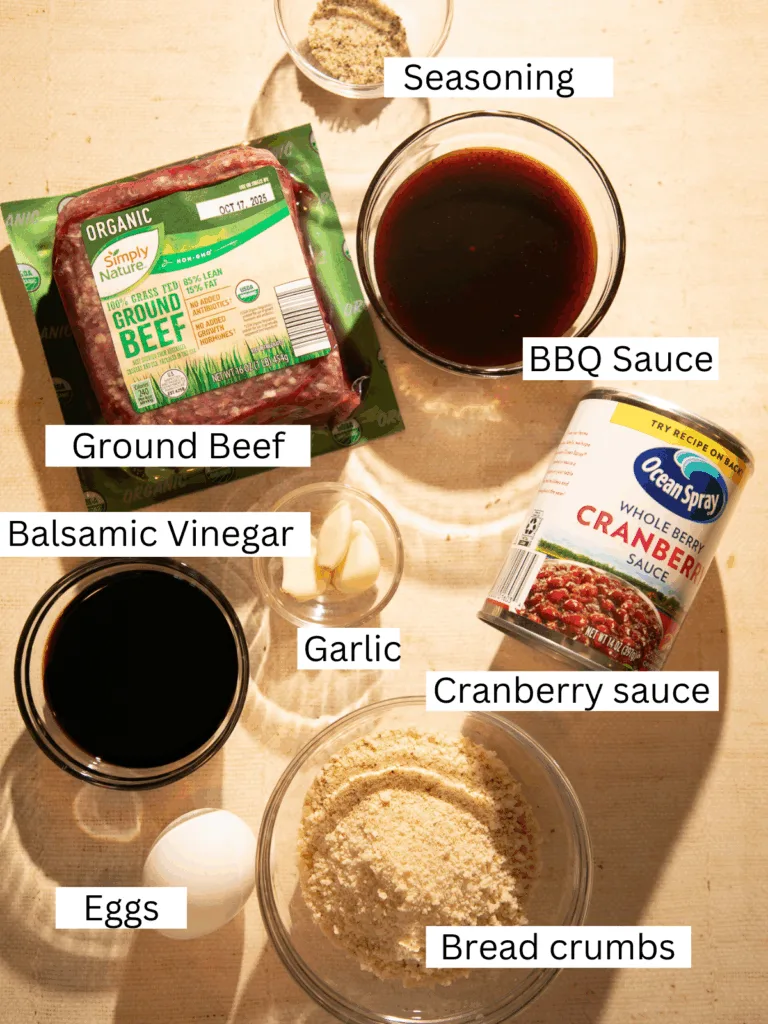 Overhead view of ground beef, cranberry sauce, BBQ sauce, breadcrumbs, eggs, garlic, balsamic vinegar, and seasonings arranged on a counter for cranberry balsamic meatballs.