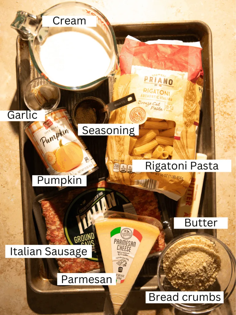 Overhead view of ingredients for brown butter pumpkin Alfredo pasta including rigatoni, Italian sausage, cream, butter, pumpkin puree, Parmesan, garlic, and breadcrumbs.