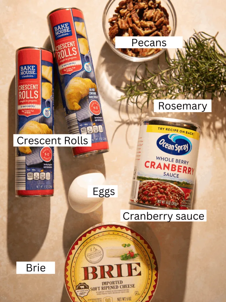 Overhead shot of ingredients for cranberry brie crescent ring including crescent rolls, brie, cranberry sauce, pecans, rosemary, and eggs.