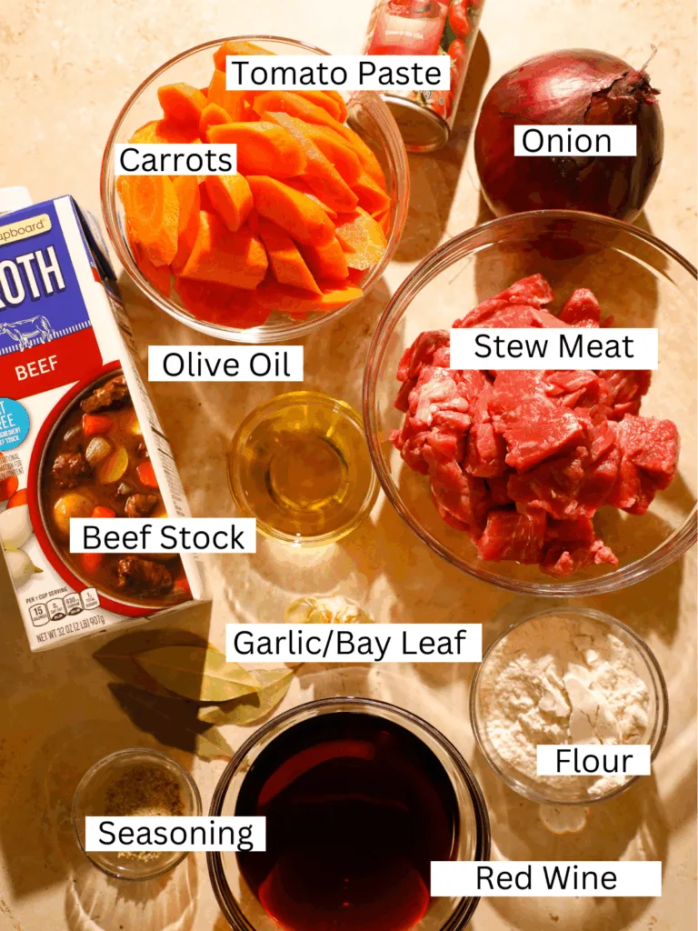 Overhead view of ingredients for Instant Pot red wine braised beef tips, including stew meat, carrots, onions, tomato paste, red wine, beef stock, and seasonings on a warm-toned surface.