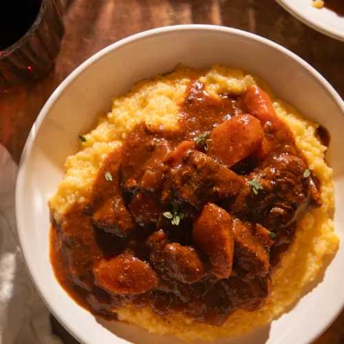Close-up bowl of Instant Pot red wine braised beef tips served over creamy polenta, topped with fresh thyme and surrounded by warm lighting.