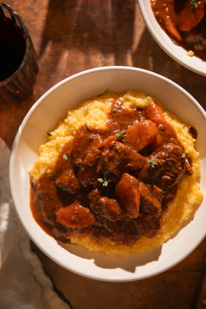 Close-up bowl of Instant Pot red wine braised beef tips served over creamy polenta, topped with fresh thyme and surrounded by warm lighting.