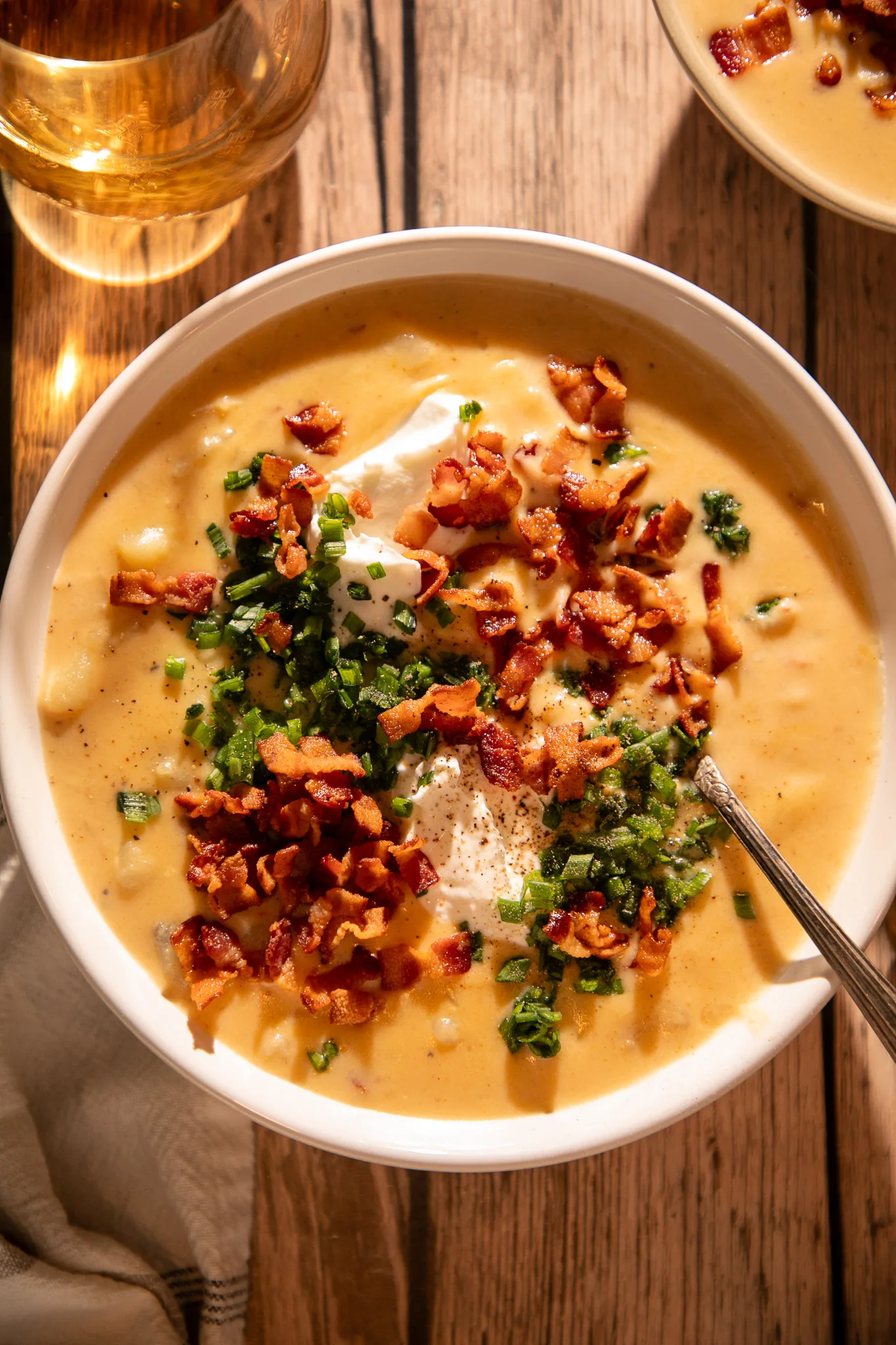 Side view of hearty potato soup with bacon, cheese, sour cream, and green onions, photographed in warm evening light on a rustic wood table.