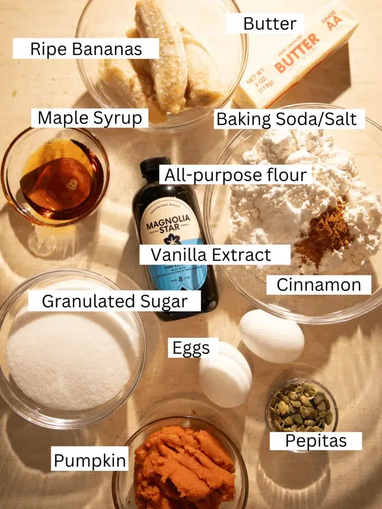 Overhead view of ingredients for spiced maple pumpkin banana bread including bananas, pumpkin puree, maple syrup, flour, butter, eggs, cinnamon, and pepitas.