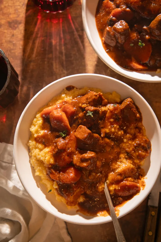 Overhead view of two bowls of tender beef tips in red wine sauce over creamy polenta with glasses of red wine beside them.