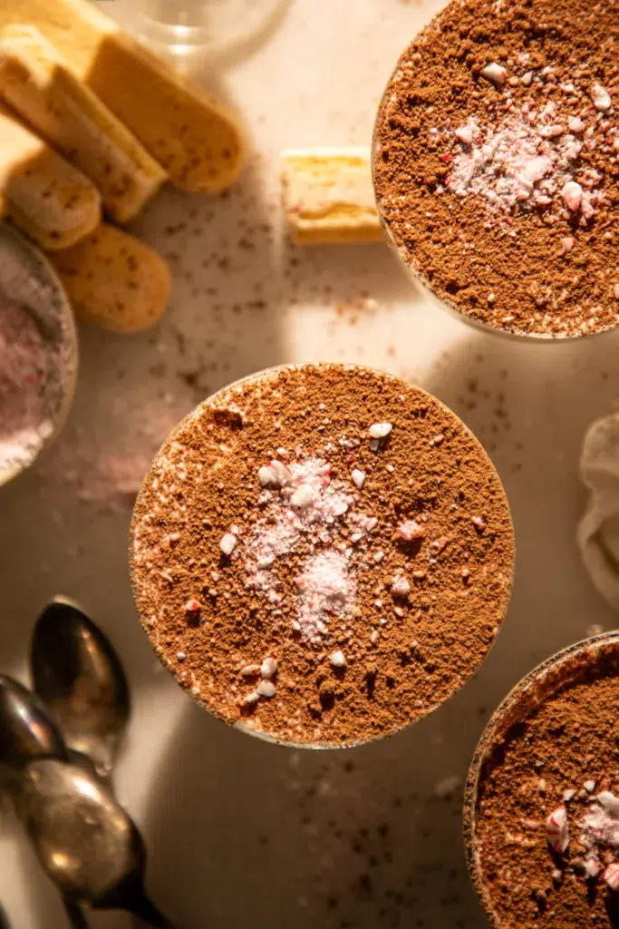 Three mini peppermint mocha tiramisu cups topped with cocoa powder and crushed peppermint pieces on a bright surface.