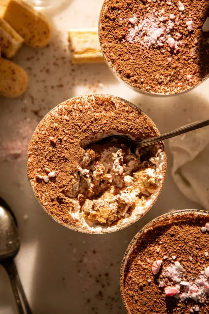 A peppermint mocha tiramisu cup dusted with cocoa powder with a spoonful scooped out, showing creamy layers and crushed peppermint.