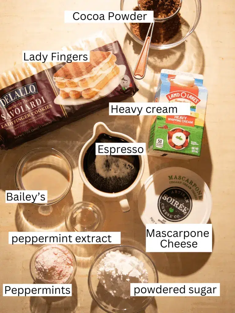 Top view of ingredients for mini peppermint mocha tiramisu cups, including ladyfingers, mascarpone, heavy cream, espresso, cocoa powder, crushed peppermints, powdered sugar, Bailey’s, and peppermint extract arranged on a warm-toned surface.