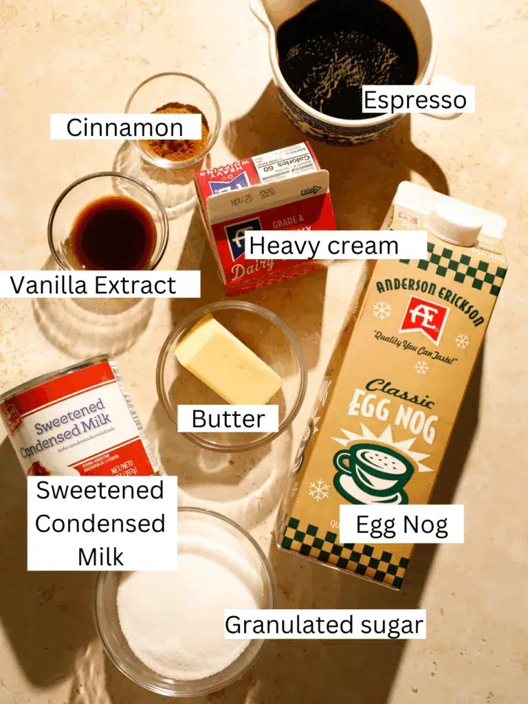 Top-down view of ingredients for eggnog affogato, including espresso, eggnog, heavy cream, cinnamon, vanilla extract, butter, sweetened condensed milk, and granulated sugar on a warm-toned surface.