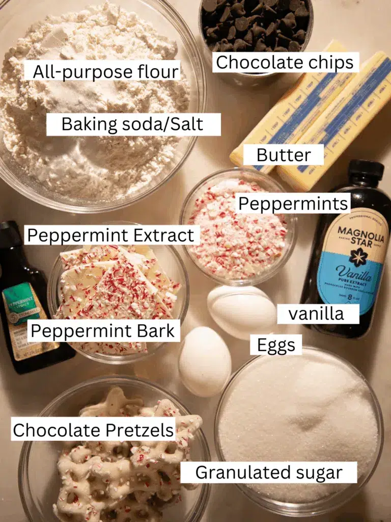 Overhead view of cookie ingredients including flour, sugar, butter, eggs, peppermint bark, chocolate chips, peppermint extract, crushed peppermints, and white chocolate pretzels arranged in small bowls.