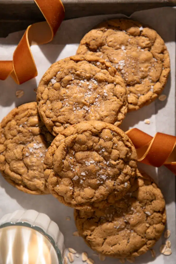 A stack of gingerbread oatmeal cookies sprinkled with sea salt on a parchment-lined tray, styled with a warm ribbon. The cookies look chewy and cozy for holiday baking.