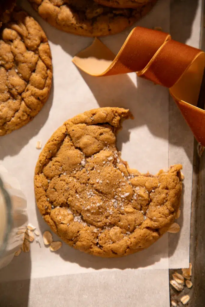 Single gingerbread oatmeal cookie with a bite taken out, sprinkled with sea salt and styled on parchment with ribbon accents.