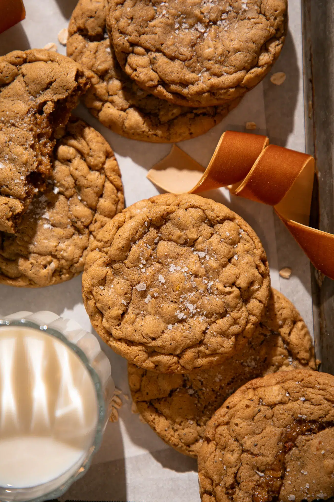 Baked brown butter gingerbread oatmeal cookies on parchment with flaky sea salt and a ribbon, served next to a glass of milk. The cookies are soft, crinkled, and golden-brown.