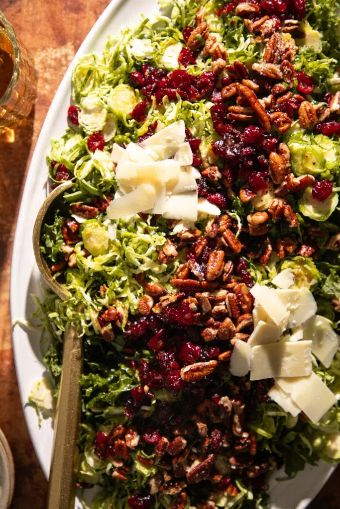 Close up of shaved Brussels sprout and kale salad topped with dried cranberries, toasted pecans, and shaved cheese on a white platter.
