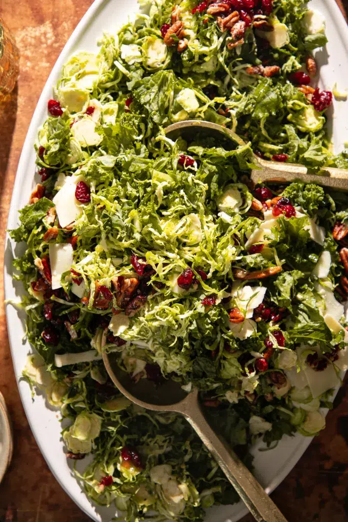 Overhead view of shaved Brussels sprouts and kale tossed with dried cranberries, toasted pecans, and shaved cheese, served with gold salad servers.