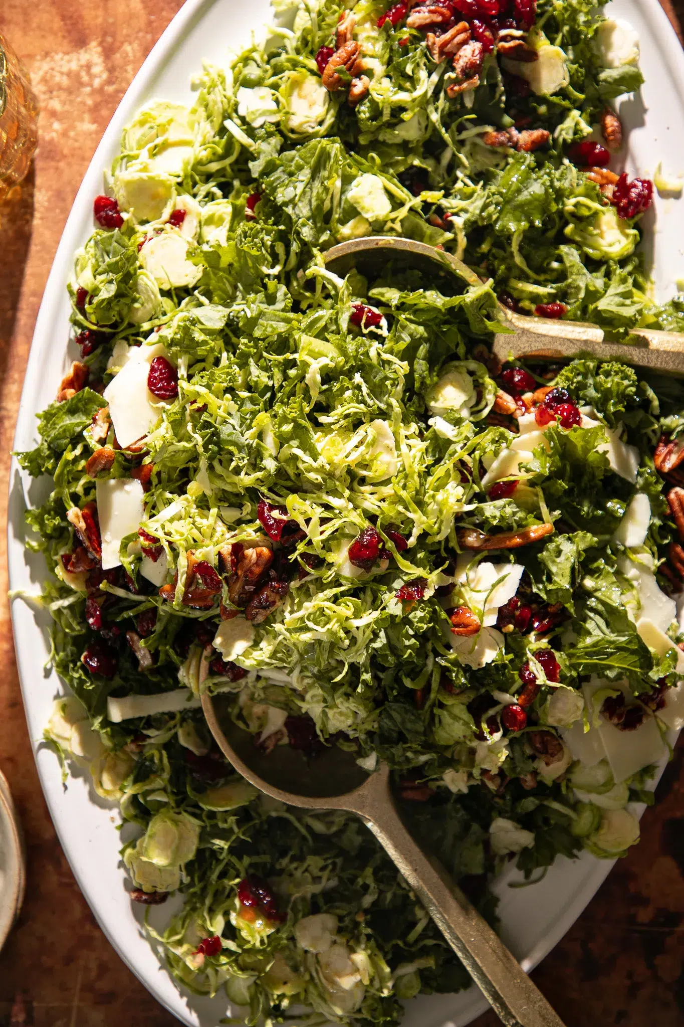 Overhead view of shaved Brussels sprouts and kale tossed with dried cranberries, toasted pecans, and shaved cheese, served with gold salad servers.