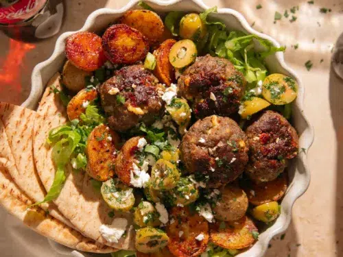 Close up of lemon feta meatballs with a golden crust, roasted baby potatoes, chopped herbs, and crumbled feta cheese.