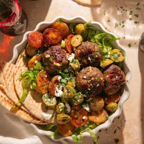 Close up of lemon feta meatballs with a golden crust, roasted baby potatoes, chopped herbs, and crumbled feta cheese.