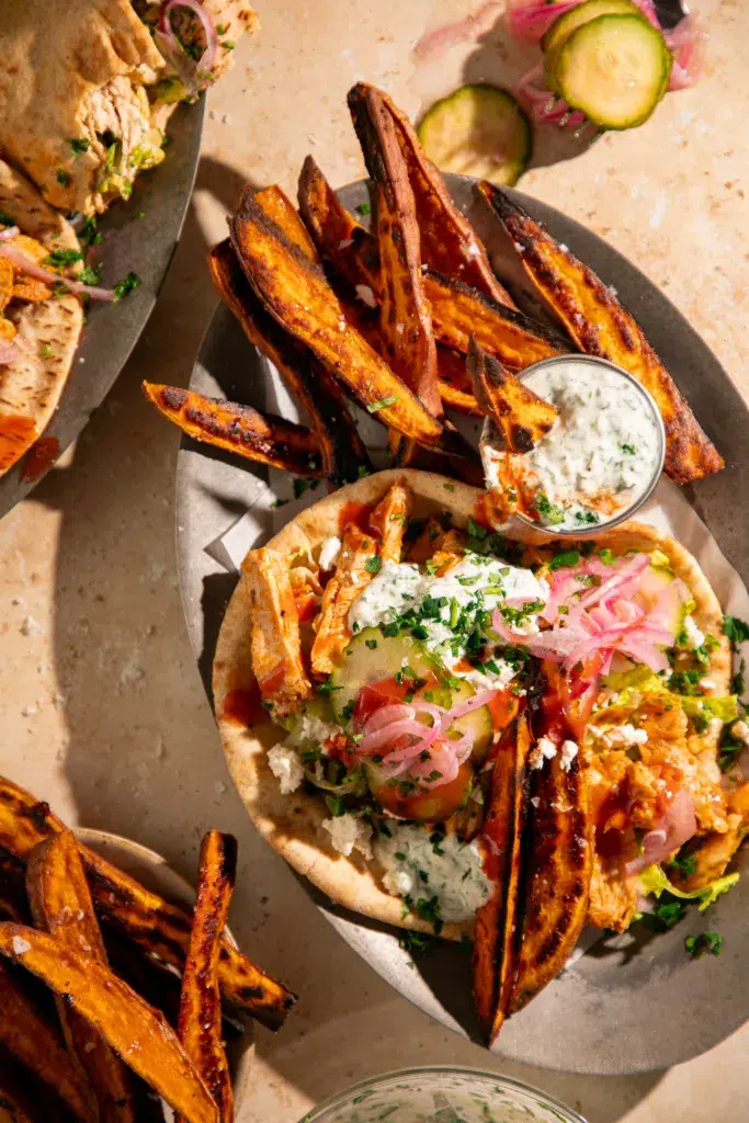 Close-up of buffalo chicken pitas topped with creamy yogurt sauce, pickled red onions, sliced cucumbers, and crumbled feta.