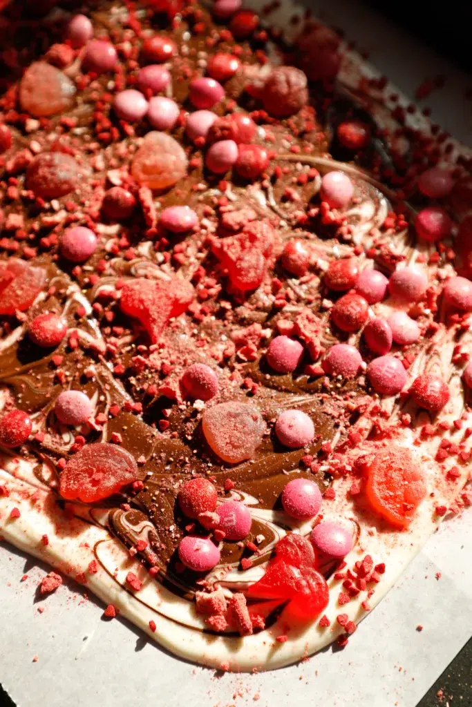 Close up of no-bake Valentine’s Day gummy bark topped with gummy hearts, pink M&Ms, crushed strawberries, and swirled milk and white chocolate.